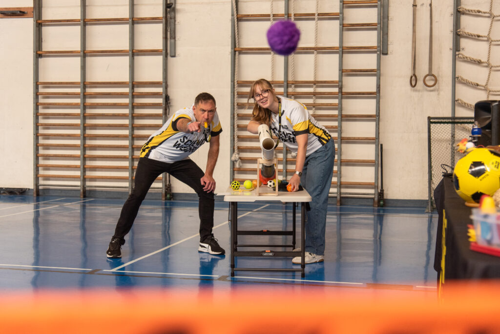 A man blows a whistle  while a woman uses a leaf-blower to send a pompom flying towards the camera.