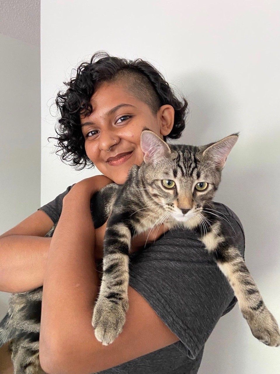 a brown-skinned agender person with short, black curly hair and a side buzz holding a cat